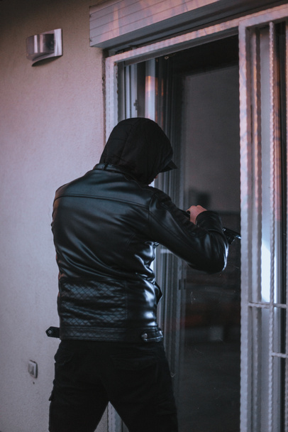Full length of hooded man using crowbar to open glass door - Valokuva, kuva