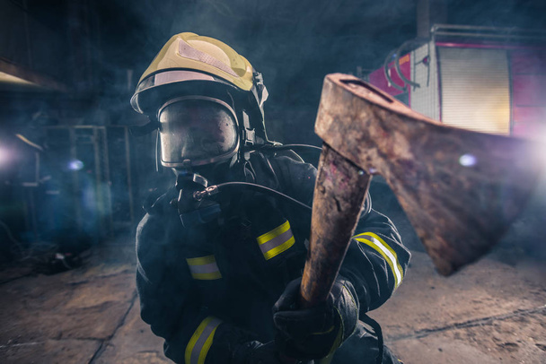 Retrato de una mujer bombero mientras sostiene un hacha y lleva una máscara de oxígeno en el interior rodeado de humo. - Foto, Imagen