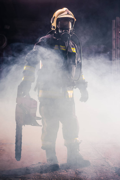 Портрет молодого пожарного, стоящего и держащего бензопилу в руках - Фото, изображение