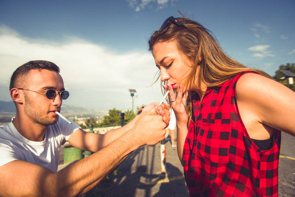 Ο τύπος ελάφρυνση ένα τσιγάρο στο ύπαιθρο κορίτσι, δαπάνες φορά μαζί σε μια ηλιόλουστη ημέρα - Φωτογραφία, εικόνα
