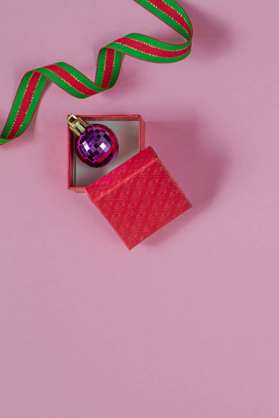 Open red gift or present box and purple christmas bubble inside with red green ribbon on pink table top view. Flat lay. Birthday, wedding or christmas concept. - Foto, afbeelding