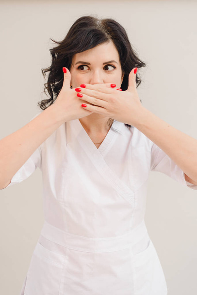 Una ragazza con un cappotto medico mostra con entrambe le mani un segno di silenzio. Un modello bruna maturo in un abito bianco cosmetologia coperto la bocca con le mani su uno sfondo beige in studio
 - Foto, immagini
