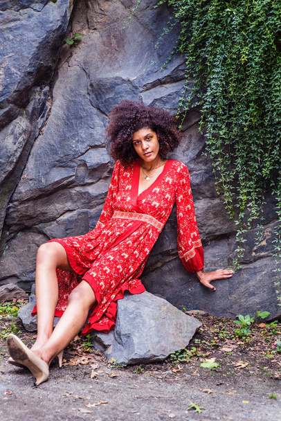 Young Mixed Race African American Woman with afro long curly hair traveling, relaxing at Central Park, New York City, wearing red flower patterned dress, high heels, stilling against rocks, thinking - Fotoğraf, Görsel