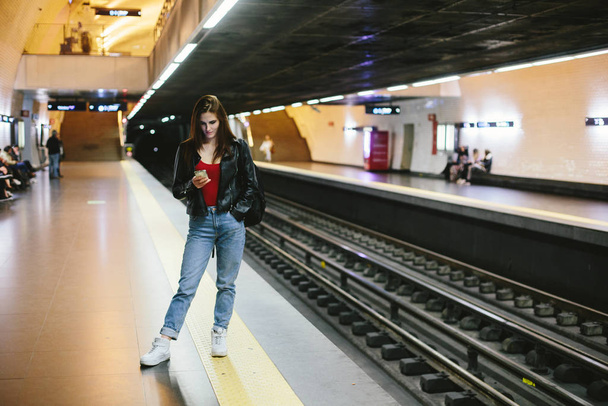 Joven hembra casual esperando el tren en la estación de metro, utilizando el teléfono móvil
 - Foto, Imagen