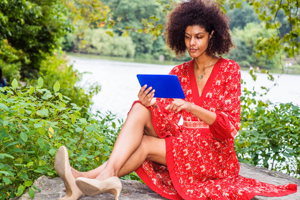 Fiatal vegyes fajú afro-amerikai nő utazik, pihentető Central Park, New York City, piros mintás ruhát visel, magassarkú, kezében kék tabletta számítógép, ül a sziklákon a tó mellett, olvasás. - Fotó, kép