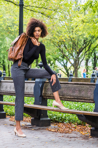 Γυναικεία μόδα στη Νέα Υόρκη. Νεαρή Αφρο-Αμερικανίδα με αφρο μακρύ σγουρό χτένισμα, ντύσιμο σε μαύρο πουλόβερ, σκούρο γκρι παντελόνι, ανοιχτόχρωμα παπούτσια, μεταφορά καφέ δερμάτινη τσάντα σε shoudde - Φωτογραφία, εικόνα