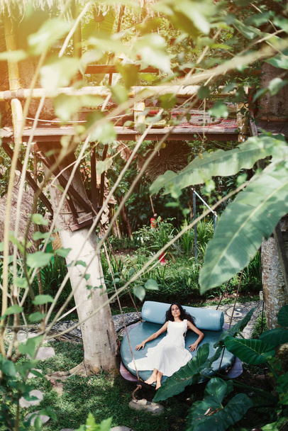 Fashion woman wearing natural white fabrics dress lying outdoor on mattress swing in jungle forest resort - Фото, зображення