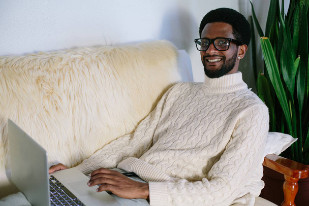 Portrait of young smiling black man wearing glasses seating on sofa using laptop at home - 写真・画像