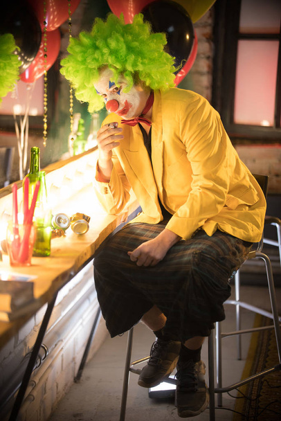 Koncept klauna - ztracený klaun ve žluté bundě a zelené paruce sedící v šatně a kouřící cigaretu po představení - Fotografie, Obrázek
