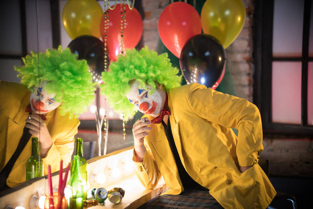 Koncepcja klauna - smutny zmęczony klaun w żółtej kurtce i zielonej peruce siedzący w garderobie i palący papierosa po występie - Zdjęcie, obraz