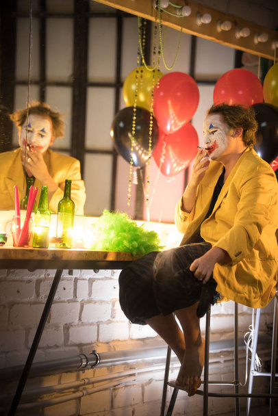 Clown concept - een trieste vermoeide man clown in gele jas roken in de kleedkamer en denken aan zijn leven - Foto, afbeelding