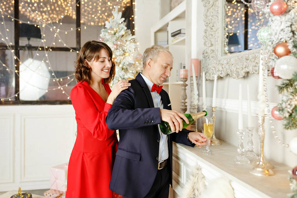 Beautiful elegant young people stand and pour champagne into glasses against the background of New Years lights in a bright studio. Merry christmas concept - Foto, afbeelding