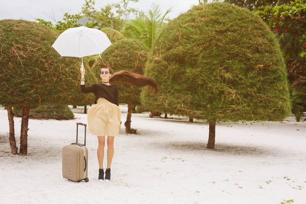 Beautiful style girl jumping with suitcase and umbrella on tropical beach - Zdjęcie, obraz