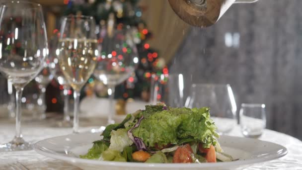 Ground black pepper from spinning pepper mill falling on salad in slow motion. Blurred Christmas lights, flashing garland in background. Xmas and New Year celebration. Seasoning dishes in restaurant - Footage, Video