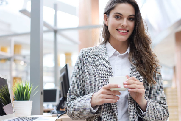 Ελκυστικός, χαμογελαστός επιχειρηματίας που στέκεται στο γραφείο με ένα φλιτζάνι καφέ. - Φωτογραφία, εικόνα