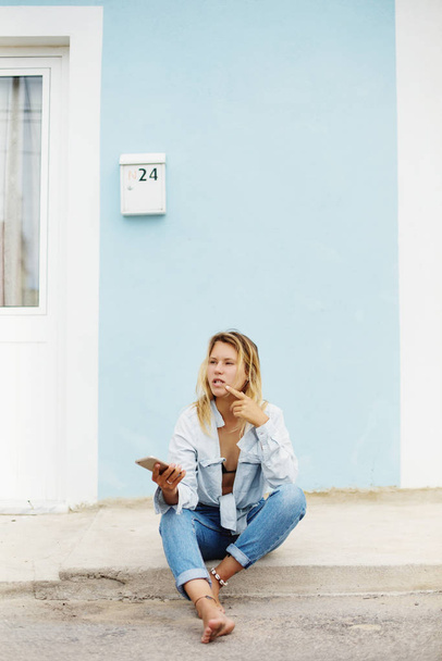 Young girl thinking and holding mobile phone, seating outdoor with blue wall on background. Space for text - Foto, afbeelding