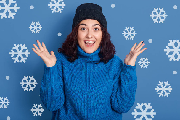 Indor shot of cute girl expressing happiness during cold winter day, adorable excited lady wearing sweater spreads hands and keeps mouth opened, posing against blue wall with snowflakes on background - Foto, imagen