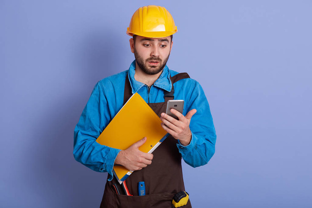 Close up portrait of young attractive engineer dresses blue shirt, brown apron and yellow helmet, holds paper folder and phone in hands, poses isolated over studio background, has shocked expression. - Foto, Imagem