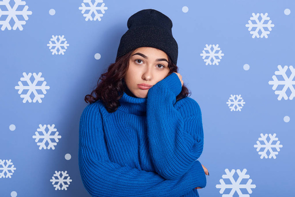Close up portrait of young brunette female standing over studio wall with snowflakes on background, keep her hand under chin and miss somebody, looking upset, wearing blue sweater and black cap. - Foto, immagini