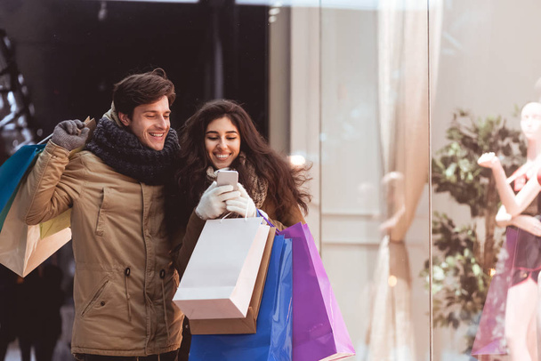 Radostné manželé pomocí mobilního telefonu vyhledávání nabízí procházky podél oken obchodů - Fotografie, Obrázek