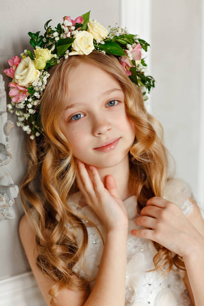 Portrait of a pretty little girl with blond hair in a pink dress is sitting in a bright studio with a wreath of fresh flowers on her head - Foto, immagini