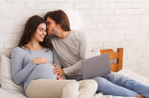 Hamile kadın ve sevgili kocası yatakta dizüstü bilgisayar kullanıyorlar. - Fotoğraf, Görsel