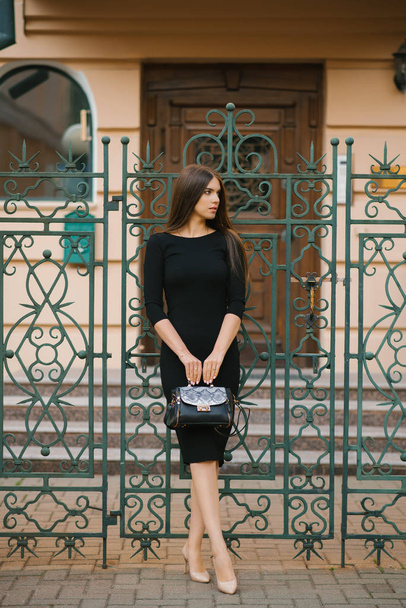 An elegant girl in a black dress holds a bag in her hands and stands near the wrought iron gate at the house - Фото, зображення