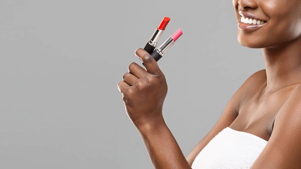 Mujer afro sonriente irreconocible sosteniendo lápiz labial rojo y rosa en la mano
 - Foto, Imagen