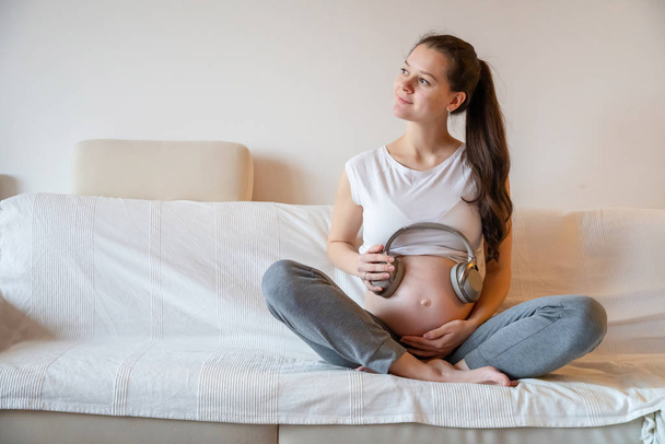 Kobieta w ciąży siedzi na białej kanapie w domu i trzyma słuchawki na brzuchu - Zdjęcie, obraz