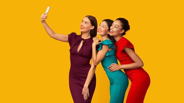 Três meninas em vestidos fazendo selfie sobre fundo amarelo, Panorama
 - Foto, Imagem