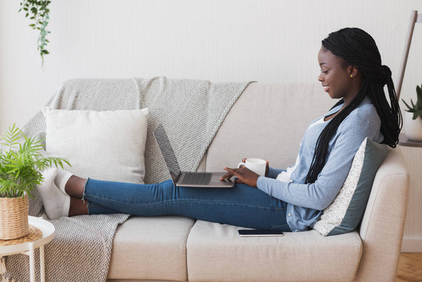 Mujer afro joven relajarse en el sofá con ordenador portátil y café
 - Foto, imagen