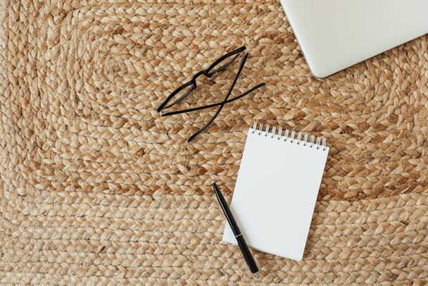 Blank sheet notebook with copy space for text, glasses, pen, laptop on wicker straw background. Flat lay, top view minimal home office desk. Work, business, education concept. - Photo, image