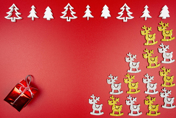Árboles de Navidad de madera blanca dispuestos en fila en la parte superior, renos dorados y plateados a la derecha y un regalo envuelto en la esquina inferior izquierda, aislado sobre un fondo rojo
. - Foto, imagen