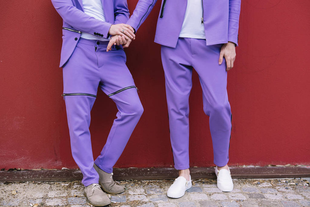 Cropped image of gay couple holding hands. Purple suits red wall on background  - Photo, Image