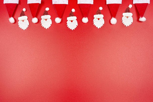 Santa Claus klobouky a Santa Claus tváře z materiálu uspořádány v řadě na vrcholu, izolované na červeném pozadí. - Fotografie, Obrázek