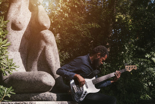 Portrait of African musician bass guitar player outdoors near the statue of a woman in the park - 写真・画像