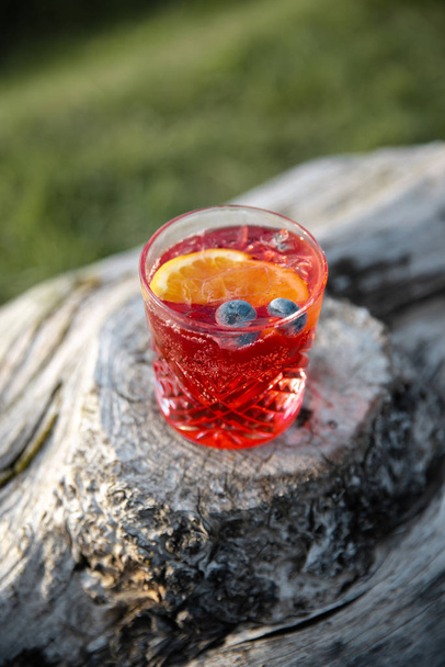 Cocktailsommer in leuchtend roter Farbe mit Blaubeere und Orange, der auf einem Holzstamm in einer Landschaft steht - Foto, Bild