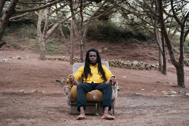 African man wearing dreadlocks and yellow shirt sitting barefoot on armchair in  forest - Photo, Image