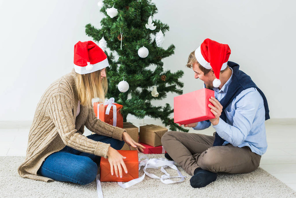 Concepto de Navidad y vacaciones - Joven pareja feliz con sombreros de santa abriendo regalos en casa
. - Foto, imagen