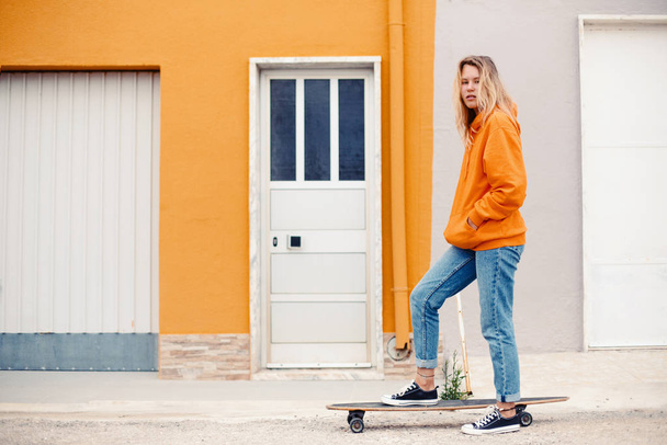 Street portrait of positive young female wearing orange hoody riding with skateboard in city - Foto, afbeelding