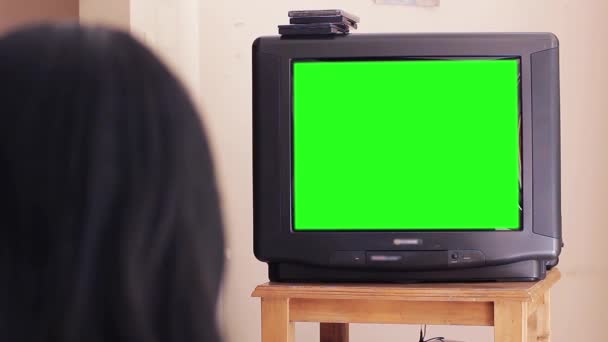 Kobieta oglądająca stary telewizor z lat 90-tych z zielonym ekranem w domu. Możesz zastąpić zielony ekran materiałem filmowym lub zdjęciem, które chcesz. Możesz to zrobić z efektem Keying w After Effects lub innym oprogramowaniu do edycji wideo (sprawdź poradniki na Youtube).  - Materiał filmowy, wideo
