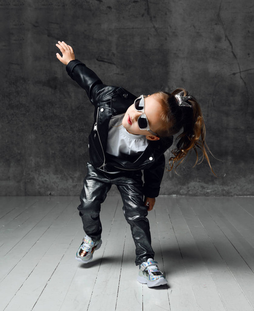 Cool frolic kid girl in leather jacket, jeans and sunglasses is having fun modern dancing playing running leaning forward - Photo, Image