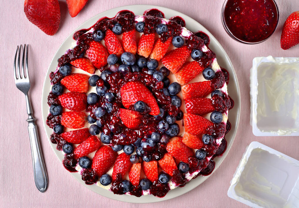 köstliche Torte mit frischen Erdbeeren, Blaubeeren und Kirschmarmelade. Gabel, leere Schalen mit Quark und Schüssel mit Marmelade. Ansicht von oben. - Foto, Bild