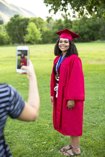 Снимок на смартфон недавней выпускницы средней школы в кепке и платье. Красивая фотография в полный рост симпатичной улыбающейся девочки африканского происхождения. - Фото, изображение