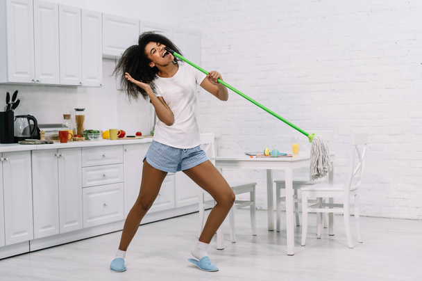 Szczęśliwa emocjonalna gospodyni tańcząca w kuchni. Piękna Afroamerykanka trzymająca mopa i śpiewająca piosenkę, bawiąca się w domu. Portret młodej, słodkiej sprzątaczki w pracy. Koncepcja prac domowych  - Zdjęcie, obraz