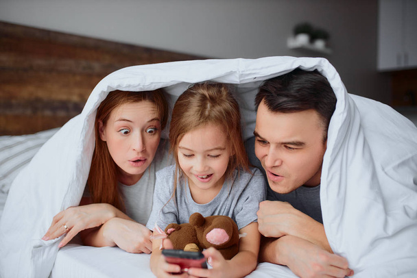 emotional parents and daughter looking at sreen of smartphone on bed - Foto, imagen