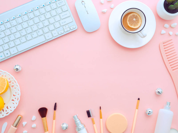 Keyboard mouse, cup of tea with slice of lemon and makeup tools and cosmetic products around it isolated on a pink background. Flat lay, top view copy space. Feminine beauty blogger workplace concept. - Photo, image