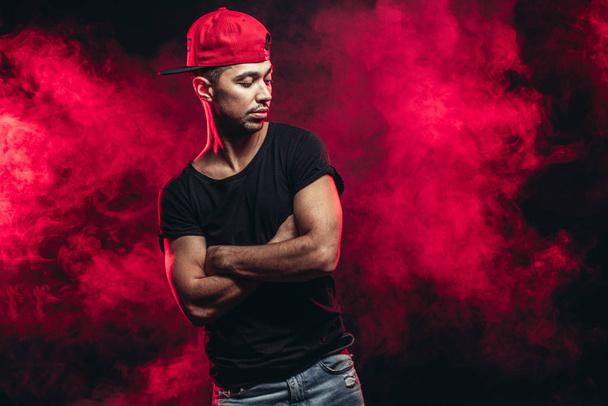 portrait of nice vigorous man wearing black t-shirt, jeans and cap isolated over red smoky background - 写真・画像
