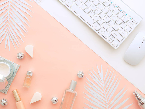 Female fashion beauty blogger home office workspace. Computer keyboard, cosmetics, perfume, cream bottles, nail polish, sponges, Christmas balls on pink, peach color background with white fern leaf - Фото, изображение