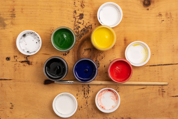 Open jars of gouache paint on a wooden background. Nearby lies a brush. View from above. - Photo, Image